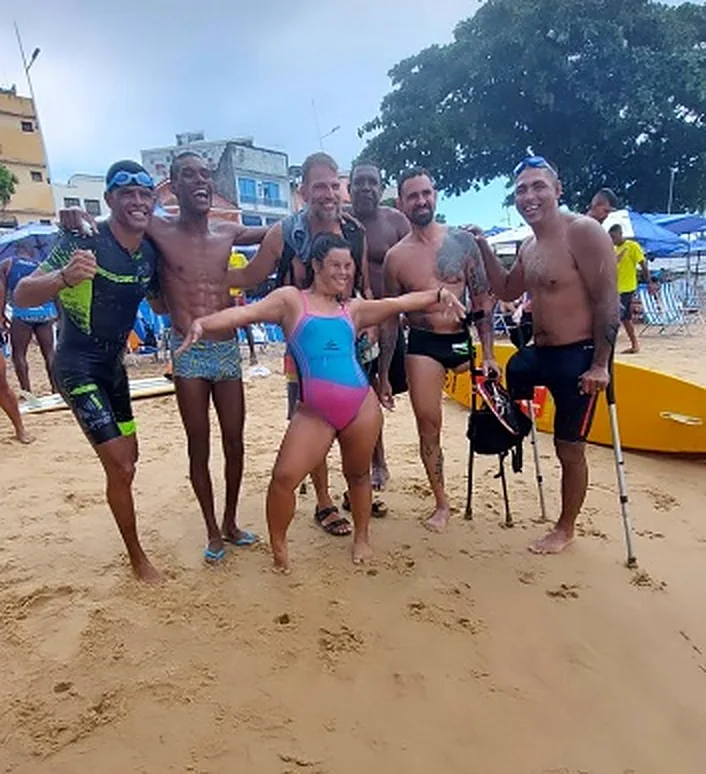 Grupo sorridente na praia, com pessoas de muletas, abraçadas para a foto.