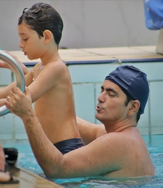 Menino e um adulto de touca, juntos na borda da piscina.