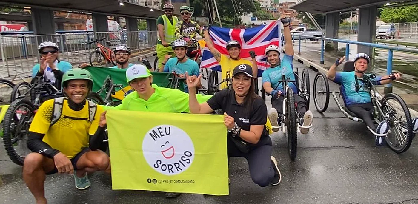 Grupo do Meu Sorriso reunido com a bandeira da associação, ao lado de handbikes e triciclos.