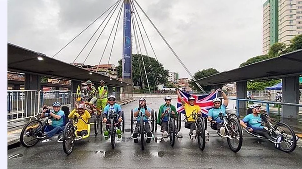 Atletas em handbikes lado a lado sob uma ponte estaiada, com bandeiras ao fundo.