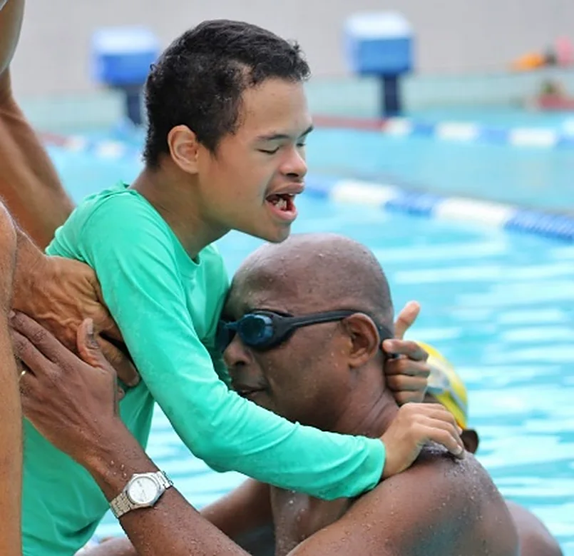 Educador abraça um jovem com deficiência dentro da piscina, os dois sorriem.