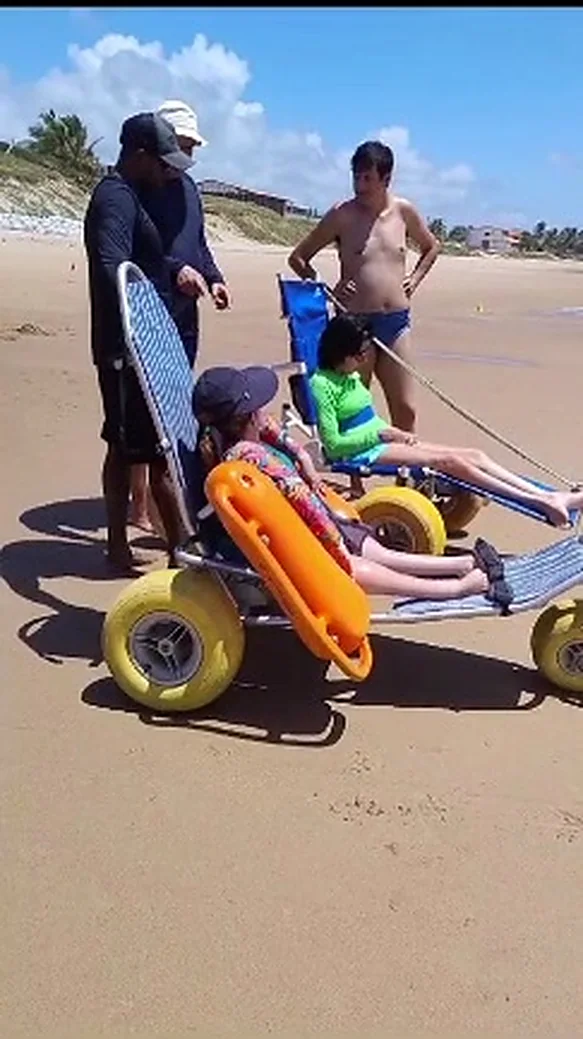 Cadeira anfíbia de rodas infláveis laranja à beira do mar, com uma pessoa sentada.