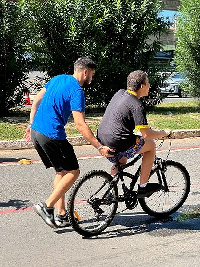 Ciclista de apoio, de camisa azul, empurra uma bicicleta comum ao lado de outro voluntário.