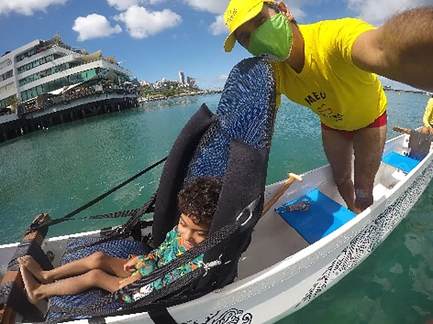 Caiaque duplo adaptado com uma criança deitada no apoio e um condutor de boné, no mar.