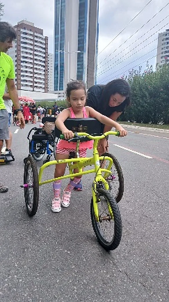 Menina pedala uma bicicleta rosa numa avenida, acompanhada por um adulto.