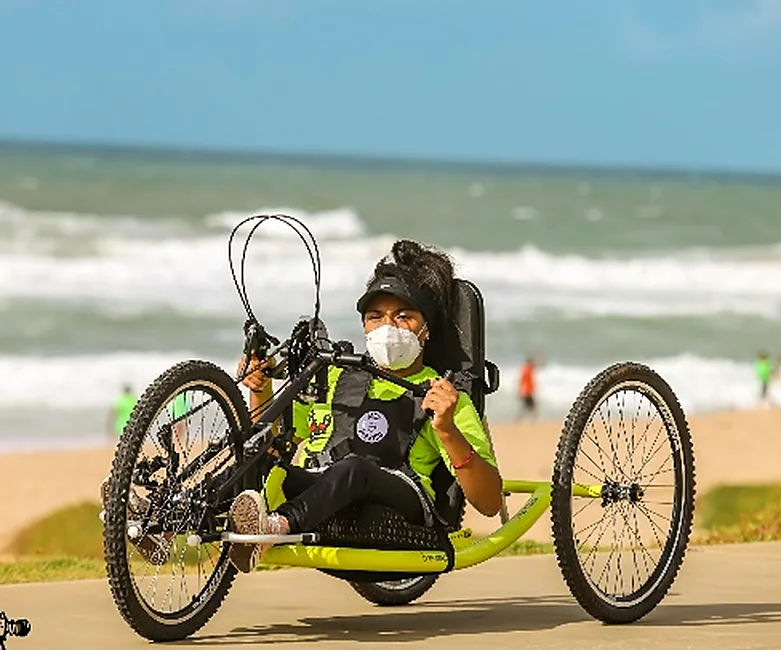 Atleta de máscara pilota uma handbike verde na areia da praia.