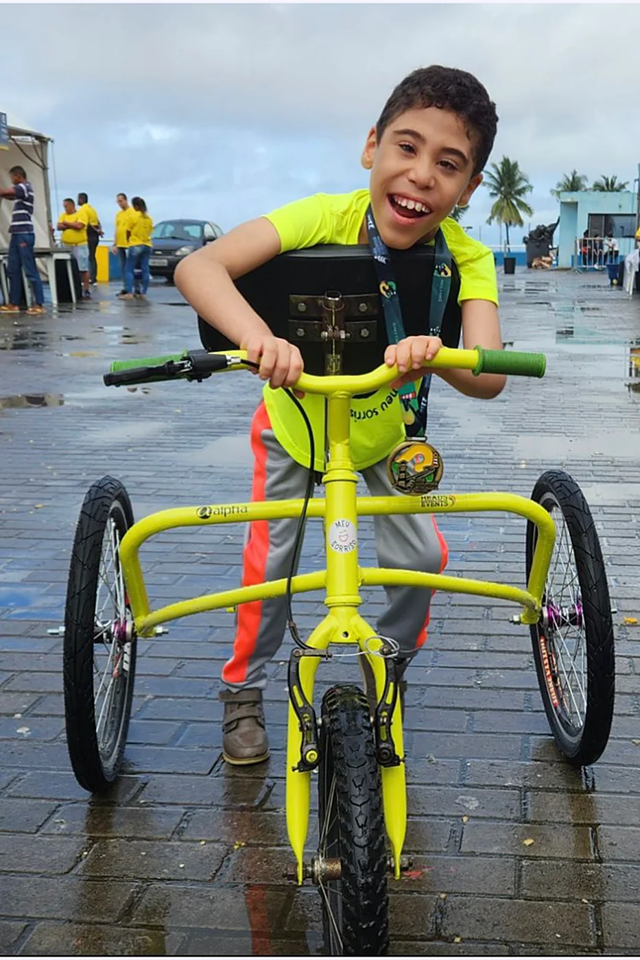 Menino sorridente pedala um triciclo amarelo adaptado, com uma medalha no peito, à beira-mar.