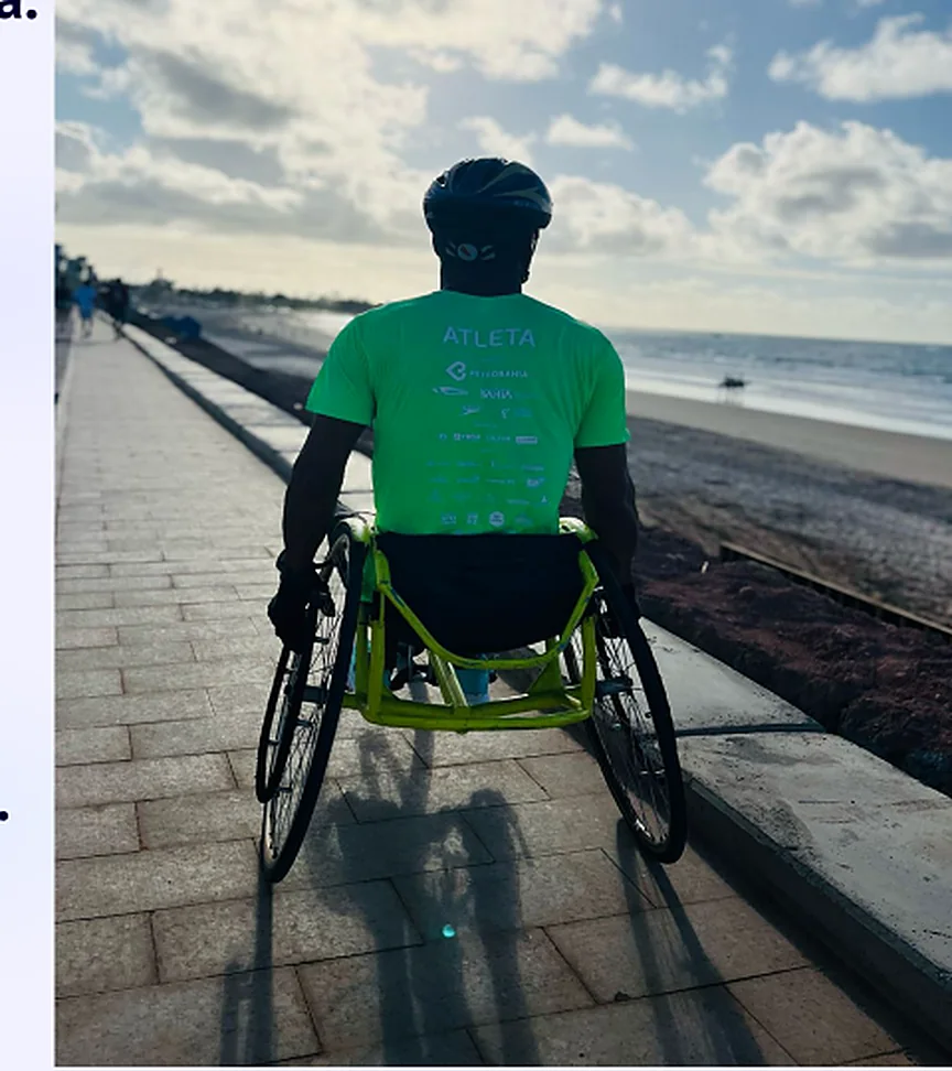 Cadeirante de costas, de capacete e camisa verde, olhando o mar ao entardecer.