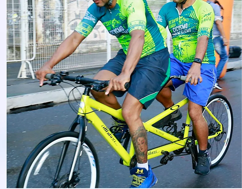 Dois ciclistas pedalam juntos numa bicicleta tandem amarela.