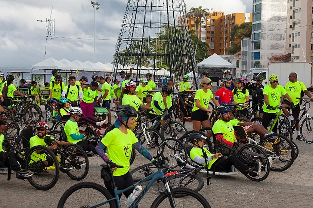 Grande grupo de ciclistas de camisa verde-limão, com triciclos e handbikes, ocupa uma avenida.