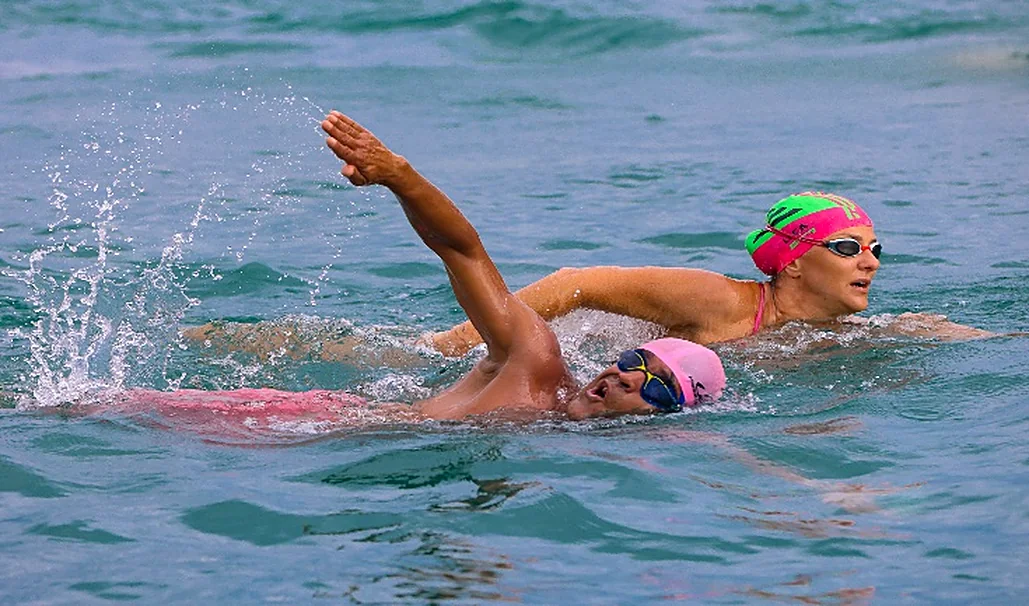 Nadador em águas abertas, de touca rosa, com braço estendido sobre a água.