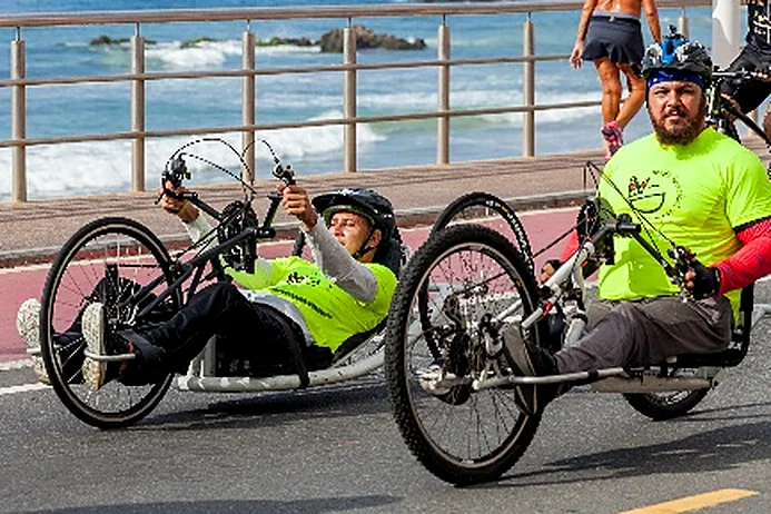 Dois atletas em handbikes lado a lado na orla, com o mar ao fundo.