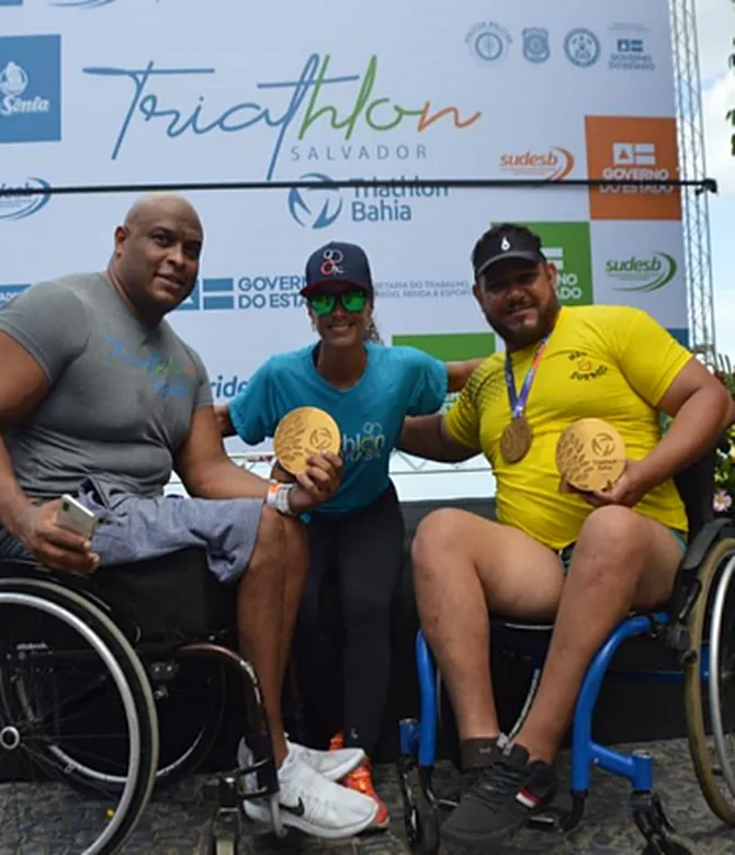 Três atletas do Meu Sorriso com medalhas, dois deles em cadeira de rodas, diante do painel do Triathlon Salvador.